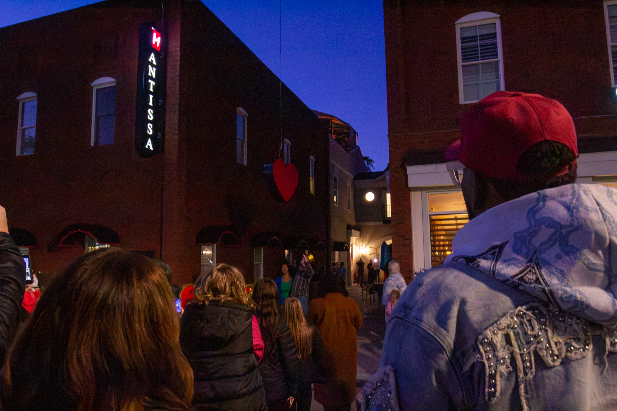 A crowd gathered at dusk as a red heart is raised beside The Mantissa's lit corner sign