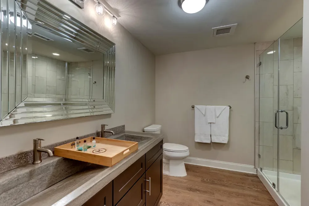 The bath of the Deluxe Dual Queen, a long mirrored double vanity with an amenity tray and the glass shower beyond.